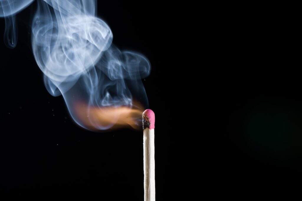 A single matchstick igniting against a black background, with smoke curling upward as the flame catches on the pink tip.