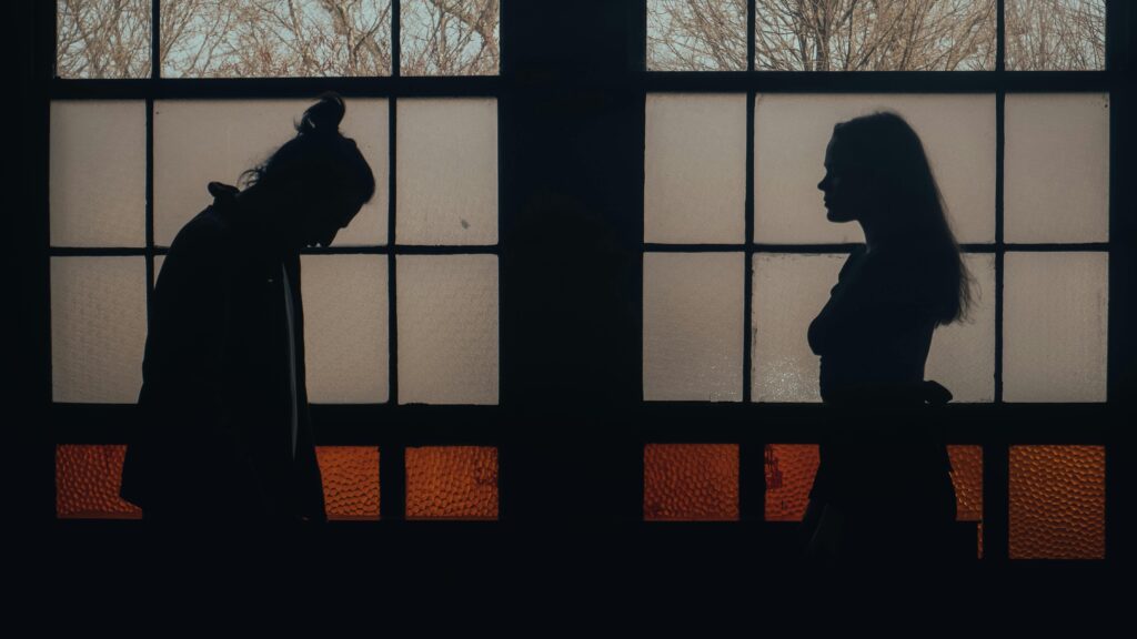Two silhouetted figures standing on opposite sides of a window frame, the man with his head lowered while the woman faces him directly.