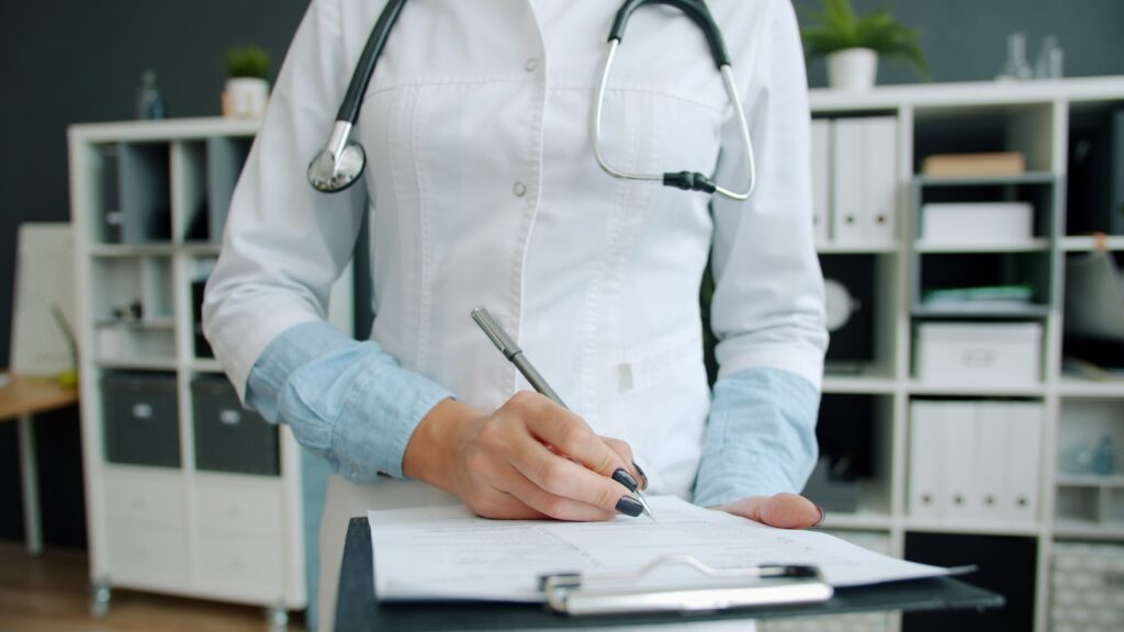 Medical professional in a white coat with a stethoscope, writing on a clipboard inside a tidy office with shelving.
