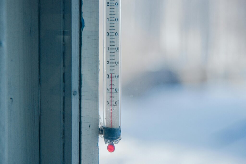 Outdoor thermometer mounted on a pale wooden window frame, reading below freezing with snow blurred in the background.