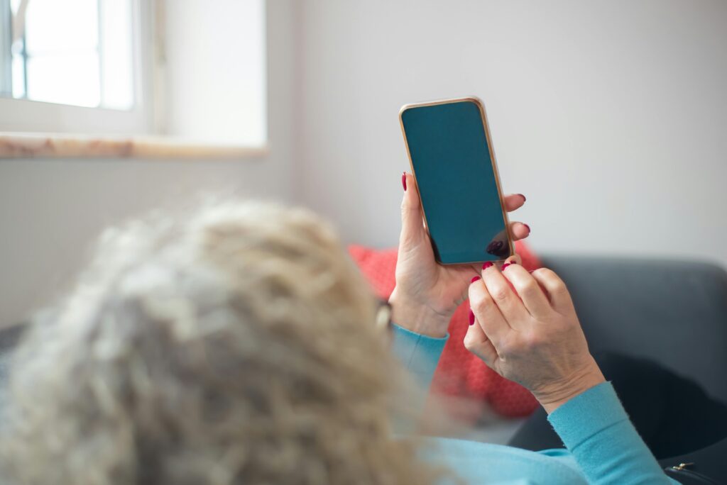 Older woman with gray curly hair seen from behind in a blue top, holding a smartphone with a blank blue screen.