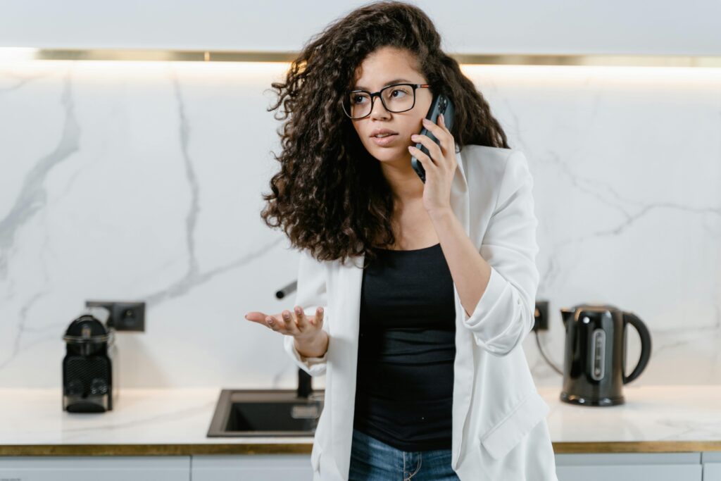 A woman with curly hair and glasses stands in a marble kitchen, holding a phone to her ear with a confused expression and her other hand raised in a questioning gesture.