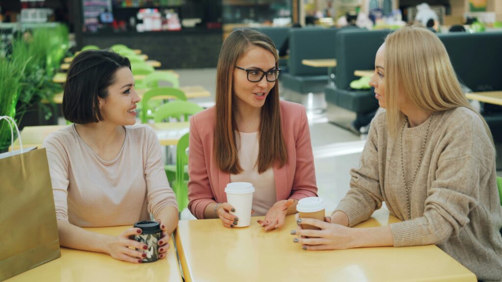 Three women sitting around a table in a cafe, chatting over takeaway coffee cups. The woman on the left has short dark hair and wears a cream jumper while holding a dark mug, the woman in the middle has long brown hair, glasses, and a pink blazer, and the woman on the right has long blonde hair and a beige knit jumper.