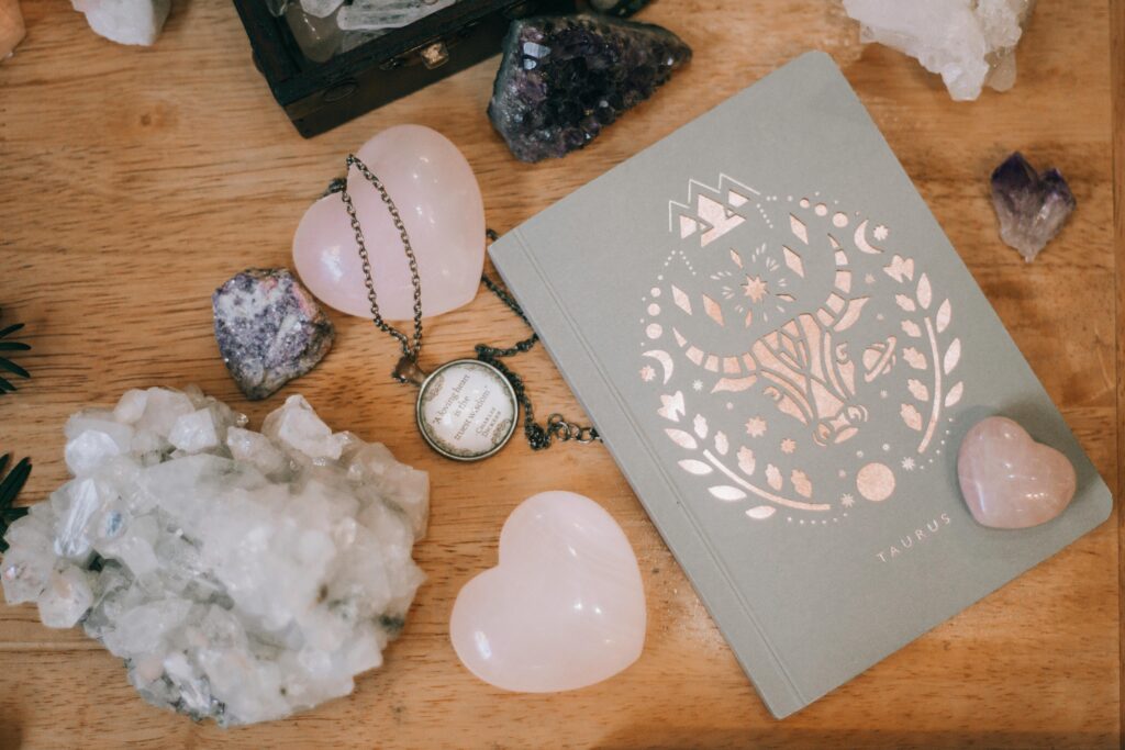 A flat lay on a wooden table featuring a sage green Taurus zodiac journal with a rose gold foil design of a bull surrounded by celestial symbols and laurel leaves. Around the journal sit various crystals including a raw white quartz cluster, a rose quartz heart, a small lilac heart-shaped stone, a dark blue sodalite piece, a purple amethyst point, and a clear quartz cluster. A vintage brass locket necklace rests on the chain beside a wooden trinket box in the top left corner.