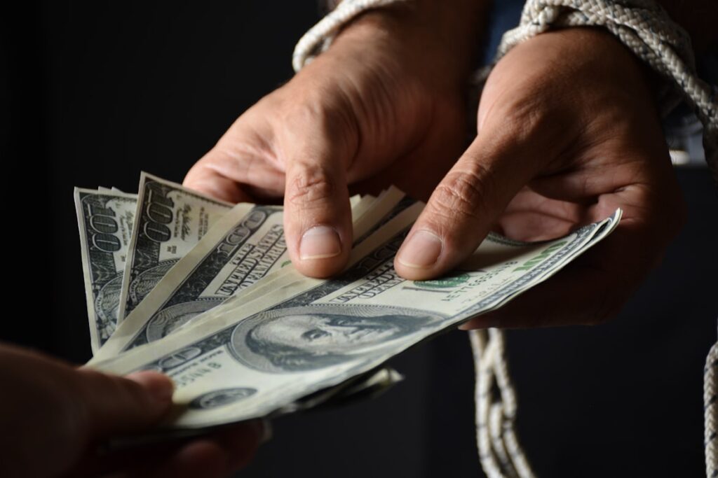 Two hands exchange a fan of hundred dollar bills against a dark background, with rope tied around one wrist suggesting entanglement or control.