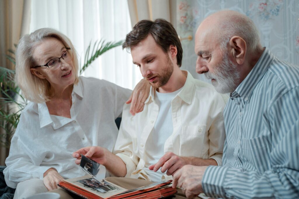 An elderly couple sits on either side of a younger man, looking together at an old photo album resting on his lap.