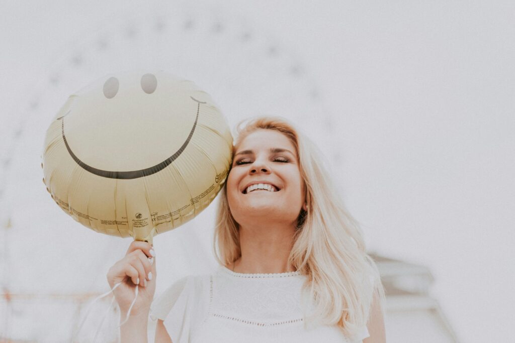 Blonde woman with eyes closed smiling widely, holding a gold smiley face balloon next to her cheek, with a blurred Ferris wheel behind her.