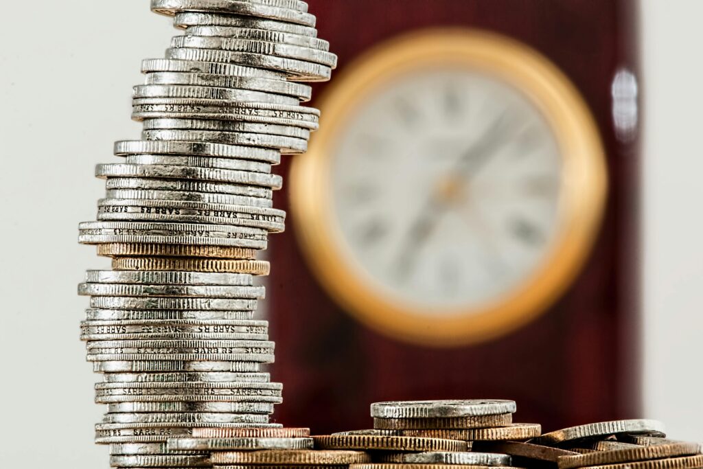 Tall stack of silver coins leaning slightly, with a blurred wooden clock face in the background.