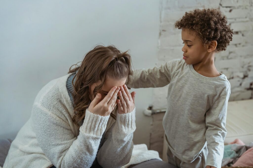 A woman sits on a bed with her face buried in her hands, while a young boy stands beside her with a comforting hand on her shoulder.