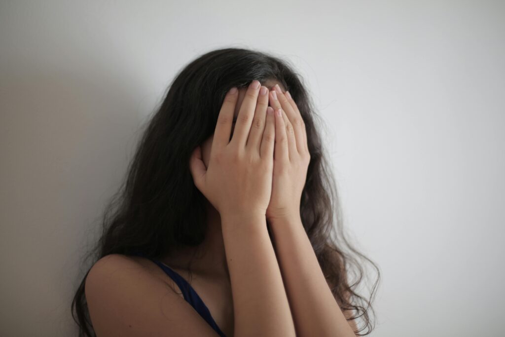 Woman with long dark hair covering her entire face with both hands, leaning against a white wall.