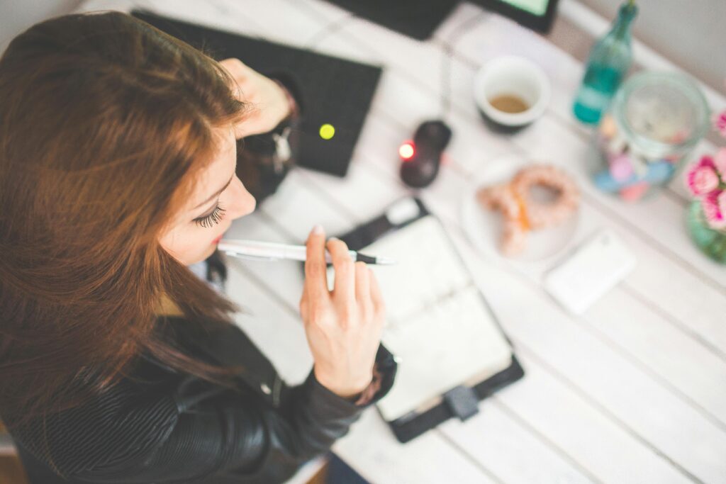 Overhead view of a woman at a cluttered desk, one hand in her hair and the other holding a pen, surrounded by a notebook, coffee, donut, and phone.