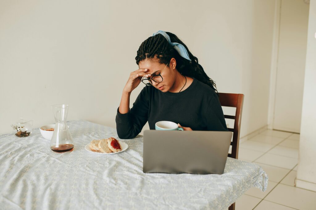 Black woman with braided hair sitting at a kitchen table in front of a laptop, holding one hand to her forehead with the other holding a mug.