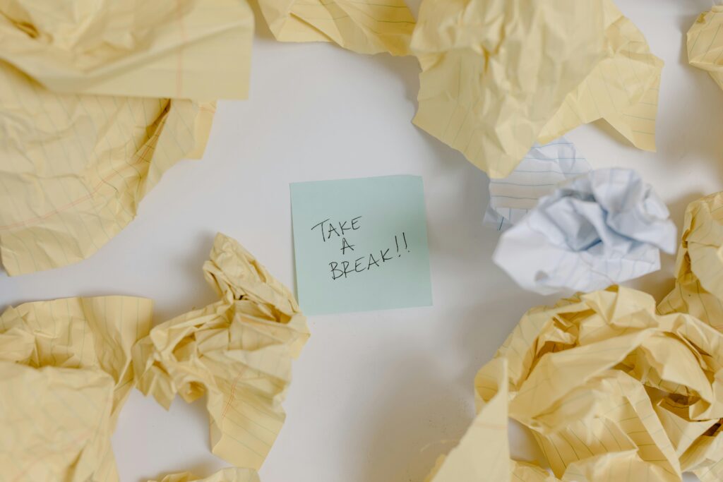 Crumpled yellow paper balls scattered around a green sticky note that reads "TAKE A BREAK!!" in black handwriting.