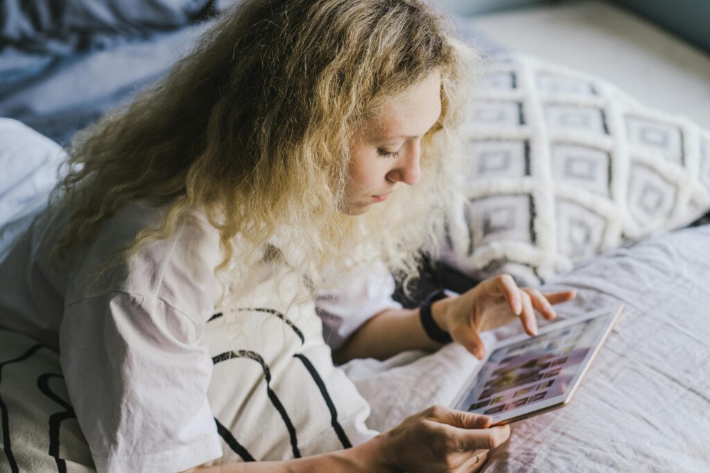 Woman with curly blonde hair lying in bed against a patterned cushion, scrolling through photos on a tablet.