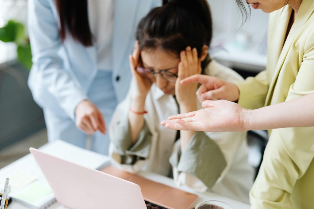 Asian woman at a desk holding her head with both hands while two colleagues stand over her, one pointing and gesturing critically.