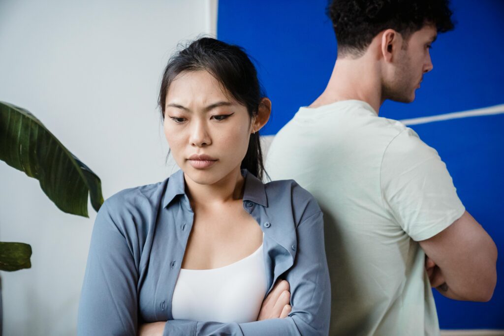 Asian woman with arms crossed standing in front of a man, both facing away from each other with tense expressions against a blue wall.