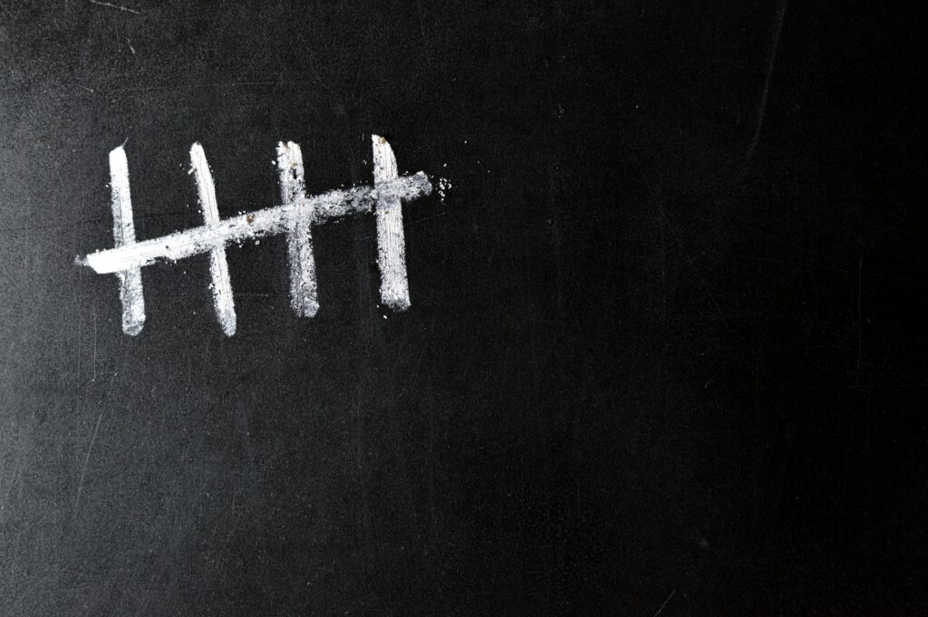 Five white chalk tally marks on a black chalkboard, with the fifth mark crossing through the first four.