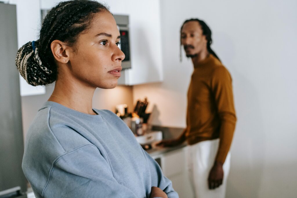 Black woman with braided hair and crossed arms standing in the foreground while a man in a mustard shirt stands blurred in a kitchen behind her.