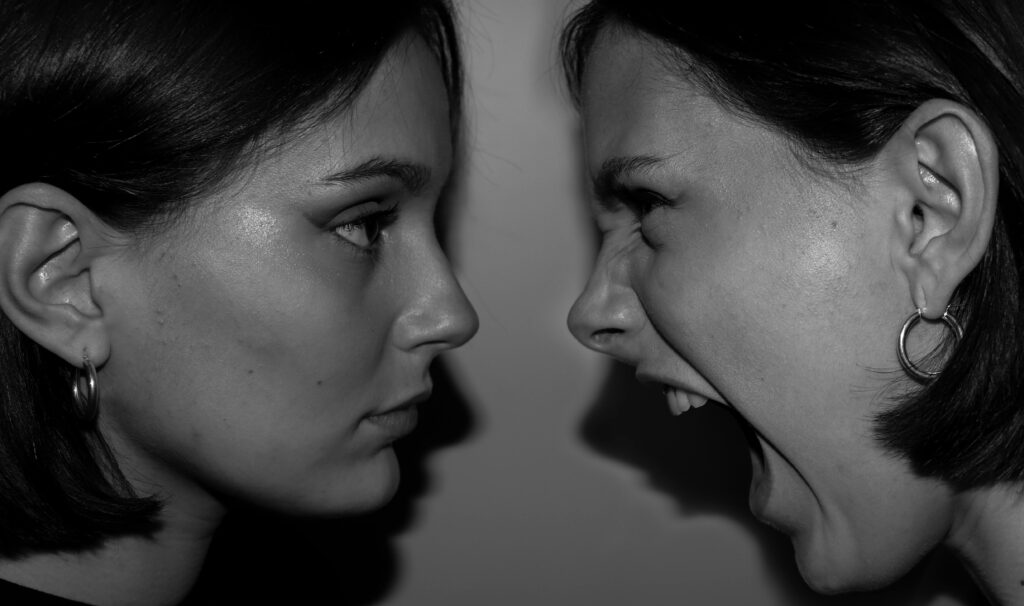 Black and white close-up of two women face to face, one calm in profile while the other screams with her mouth wide open.