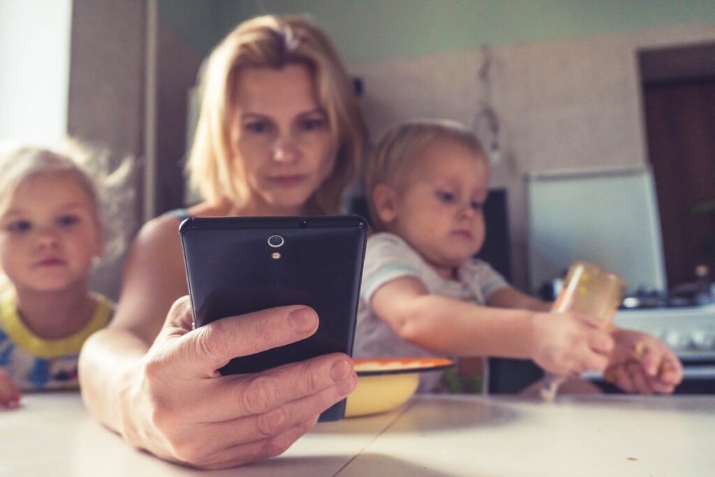 Mother in a kitchen scrolling on her phone while her toddler and young child sit at the counter beside her.