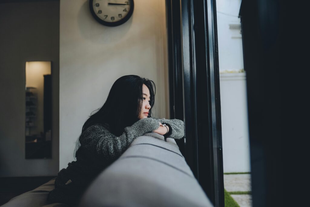 Woman with long dark hair in a grey sweater leaning on the back of a couch, gazing out a window with a wall clock visible in the background.