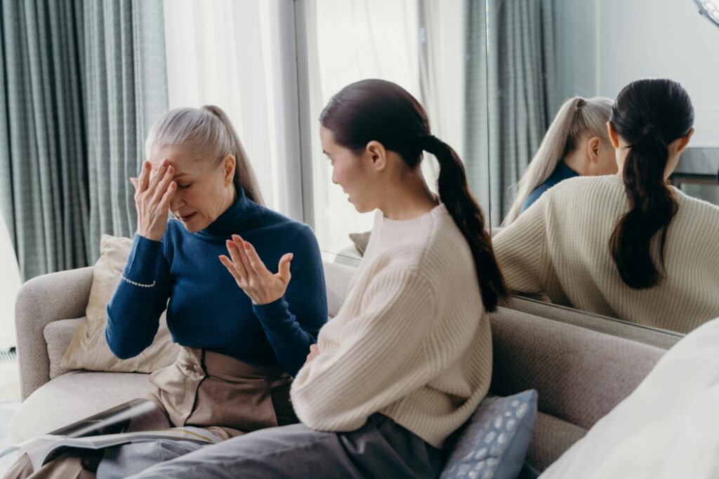 Two women sit on a couch in conversation, with the older woman holding her forehead in frustration while the younger woman gestures as she speaks.