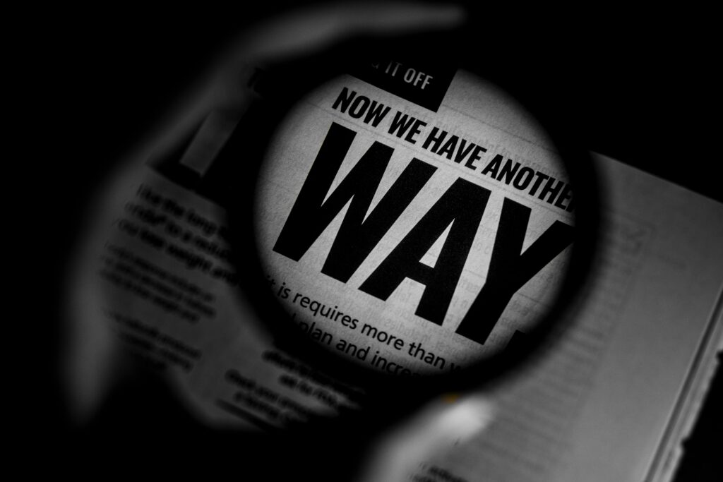Black and white photo of a magnifying glass held over newspaper text, zooming in on bold lettering that reads "NOW WE HAVE ANOTHER WAY."