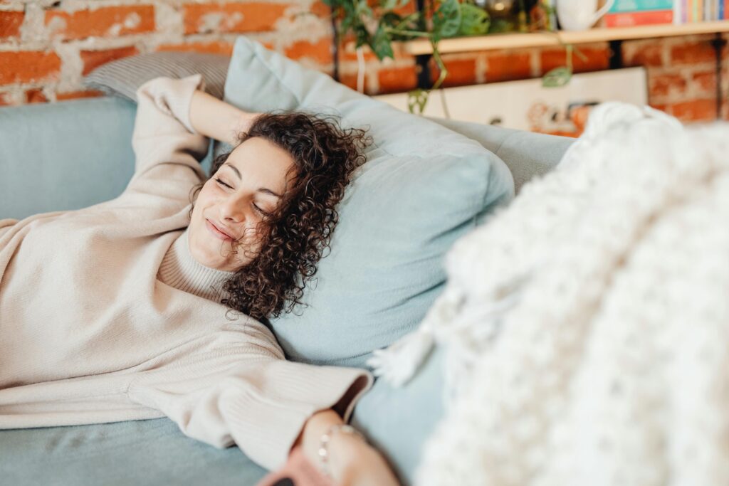 Woman with curly brown hair lying back on a blue couch with her eyes closed and a soft smile, one arm tucked behind her head, with an exposed brick wall in the background.