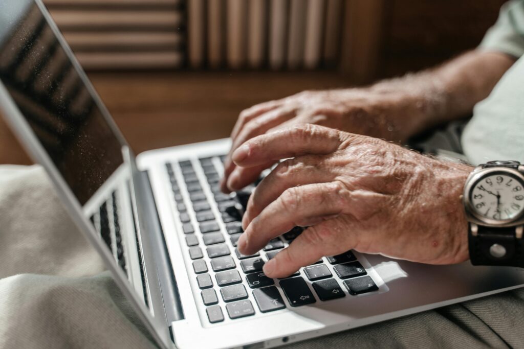 Older man's weathered hands typing on a silver laptop balanced on his lap, a leather-strapped wristwatch visible on his wrist.