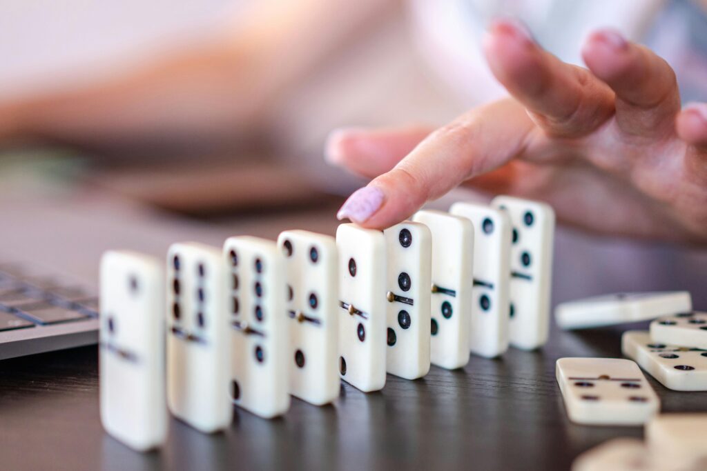 Close-up of a row of white dominoes mid-fall, with a finger tipping the first piece into the chain.