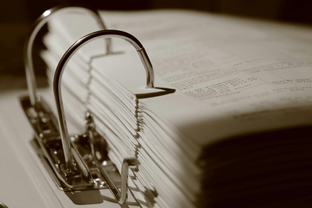 Close-up of an open binder stuffed with filed paperwork, metal rings arching over stacked documents, shown in warm sepia tones.