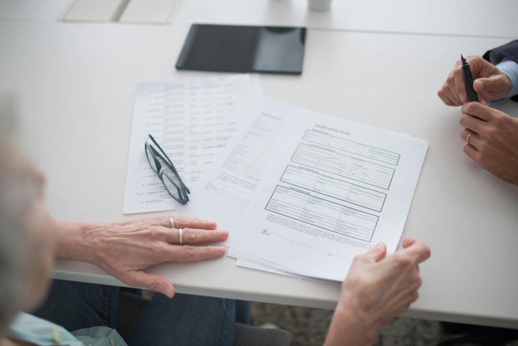 Older person's hands resting on an application form across a white table, reading glasses to the side, another person holding a pen opposite them.