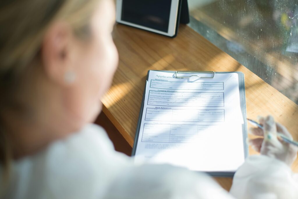 Woman seen from behind filling out an application form on a clipboard, with a tablet and sunlight streaming across the wooden desk.