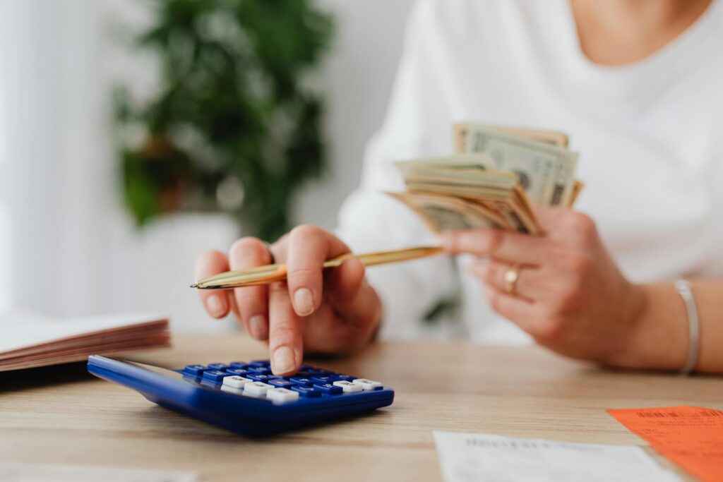 Close-up of hands at a desk, one pressing buttons on a blue calculator while the other holds a fanned stack of dollar bills.