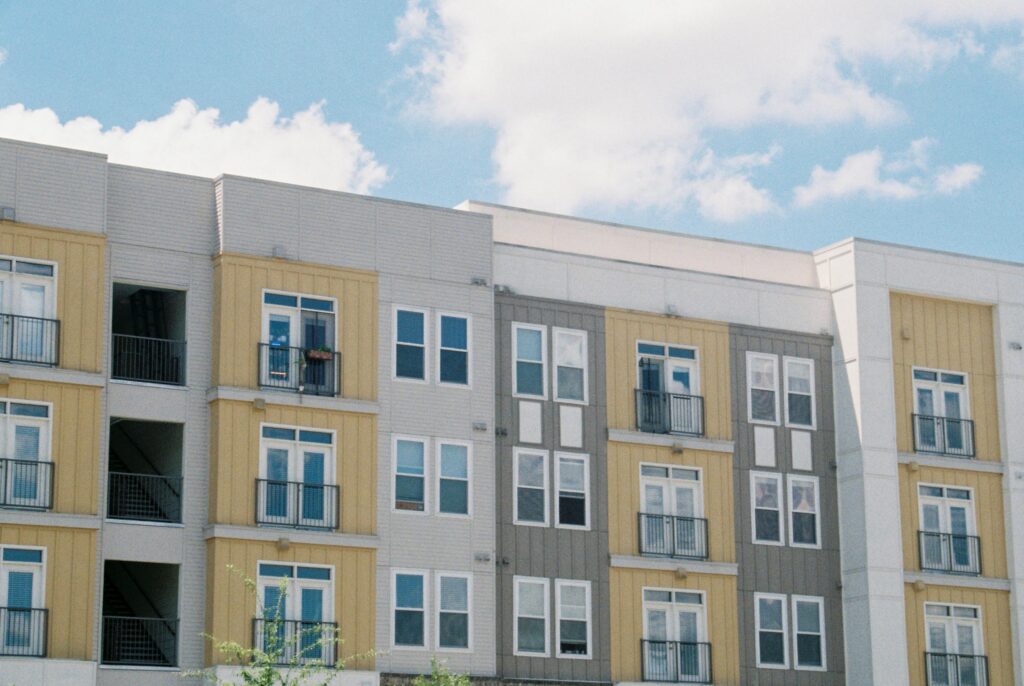 Modern apartment building exterior with yellow and gray siding, multiple floors with small balconies, photographed against a blue cloudy sky.