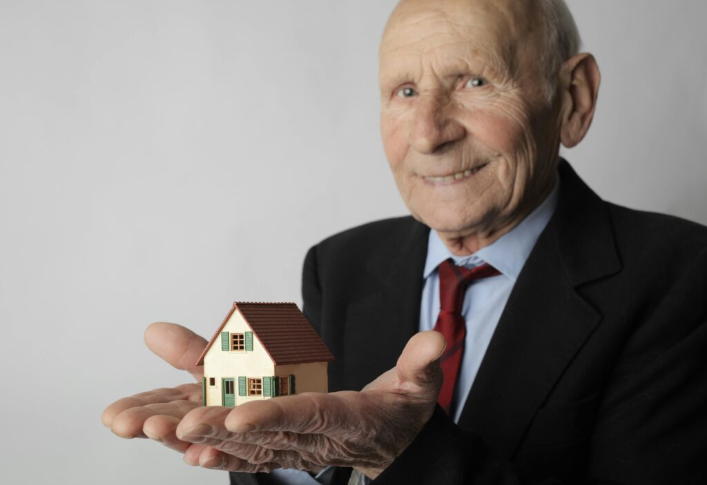 Smiling elderly man in a dark suit and red tie, holding a small model house on his outstretched palms.