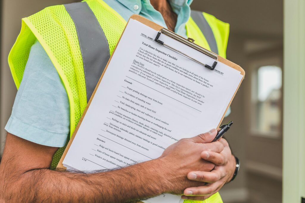 Construction worker in a yellow safety vest holding a home inspection checklist on a clipboard against their chest.