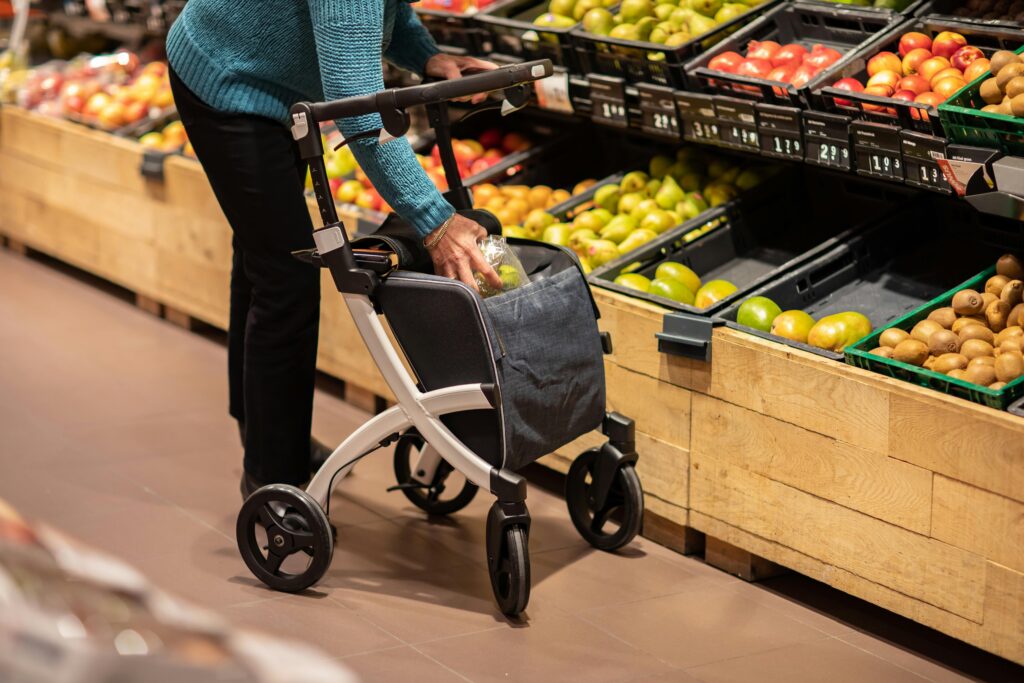 Older person using a rolling walker with a storage bag, reaching for fresh produce in a grocery store's fruit section.