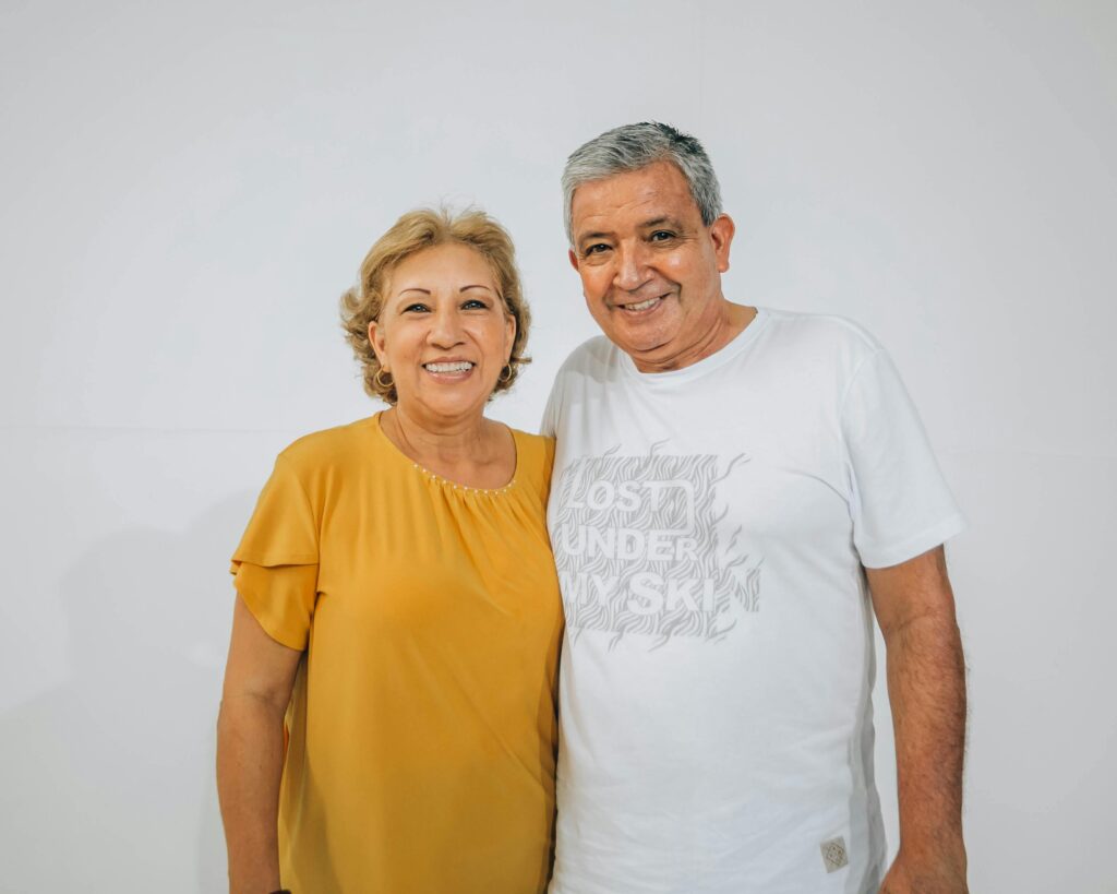 Smiling older couple posed against a plain white wall, woman in a yellow blouse with short blonde hair, man in a white graphic t-shirt with his arm around her shoulder.