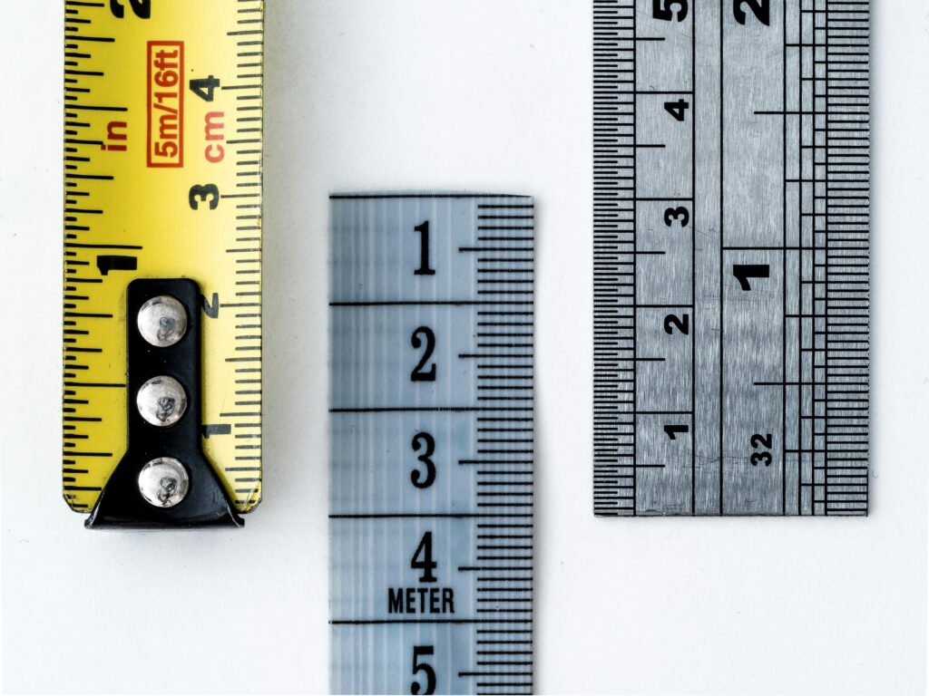 Overhead view of three different measuring tools laid side by side on a white surface, a yellow tape measure showing inches and centimeters, a clear plastic ruler marked in meters, and a metal ruler with fine gradations.