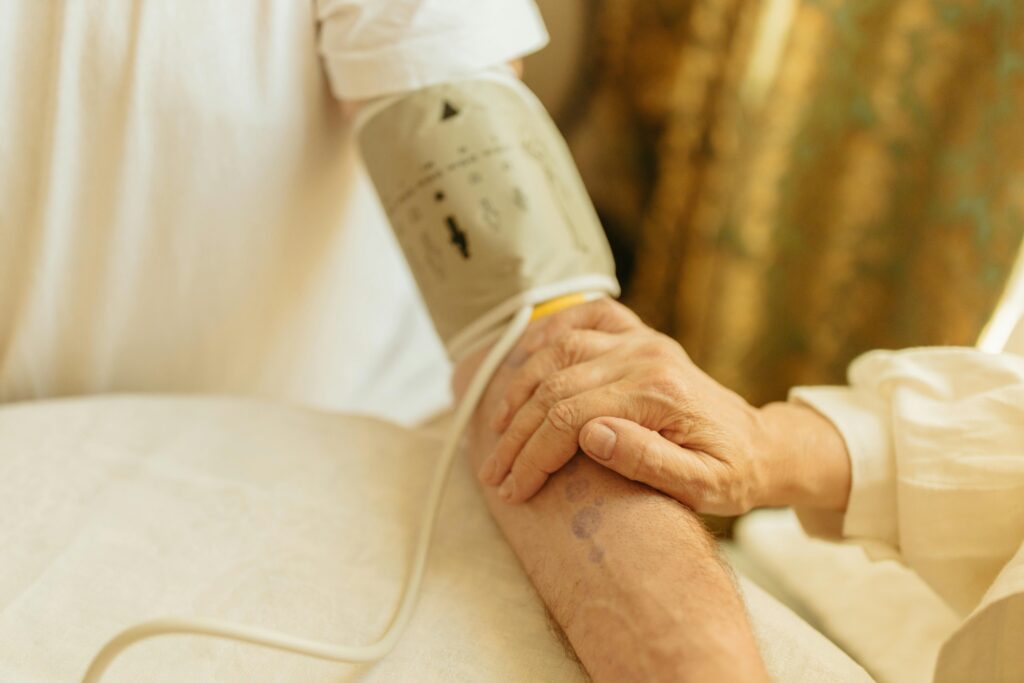 Elderly patient's arm fitted with a blood pressure cuff resting on a bed, with a caregiver's hand placed gently over their arm.