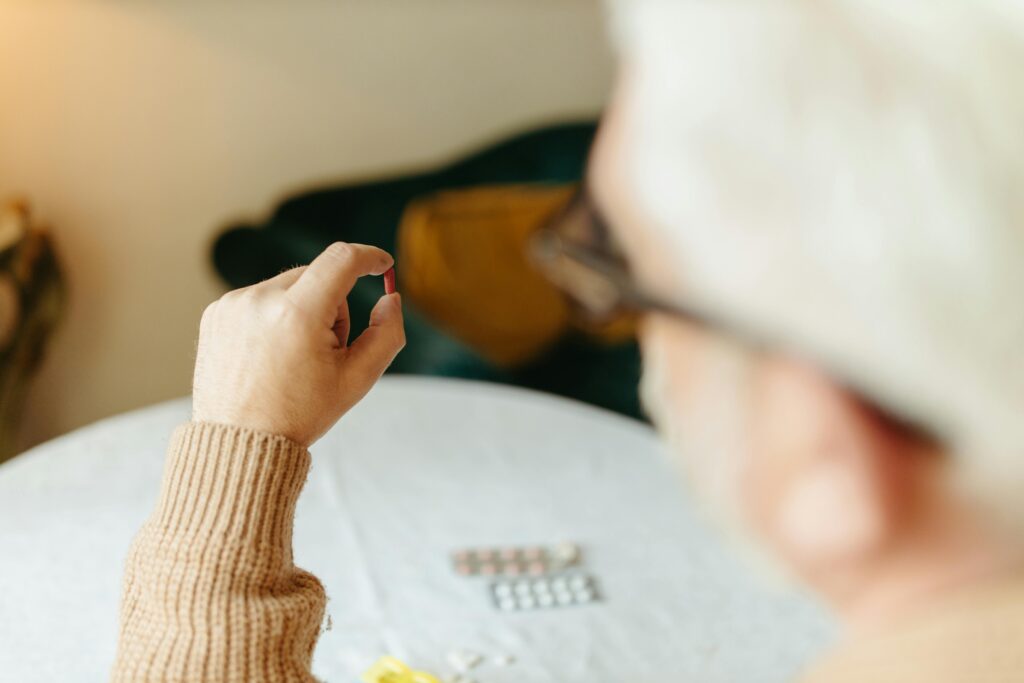 Older person with gray hair and glasses seen from behind, holding a single pill between their fingers with more pills spread on the table in front of them.