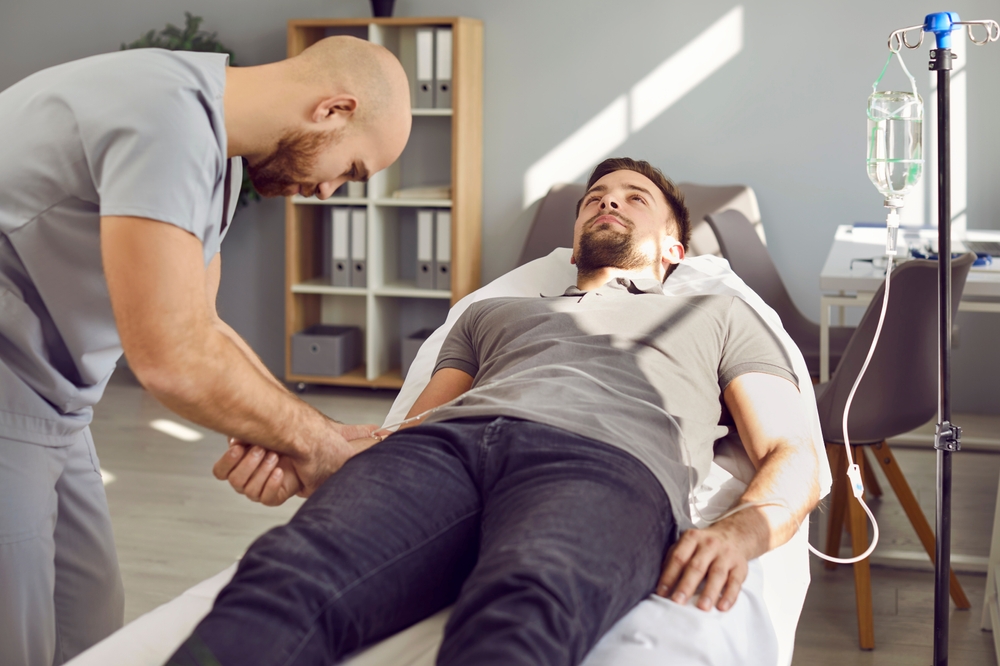 Staff nurse gives intravenous vitamin infusion to young male patient. Professional personnel puts medicine IV line catheter in vein of weak adult man lying on bed in hospital ward or medical office