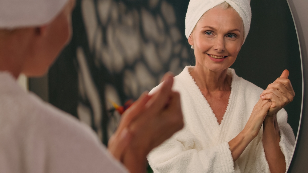 Caucasian old mature 50s middle-aged woman looking at mirror in bathroom apply cream on dry wrinkled hands body lotion product cosmetic anti-aging lifting moisturizer skin care bodycare beauty routine