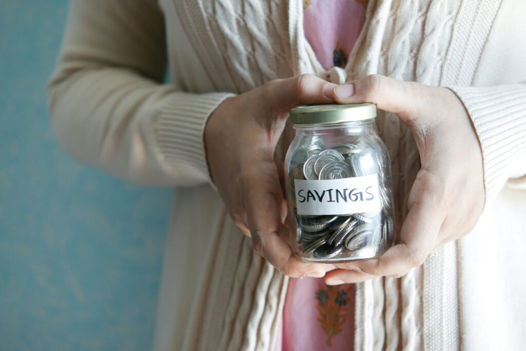 Person in a cream cardigan cupping a glass mason jar filled with coins, labeled "SAVINGS" in handwritten letters.