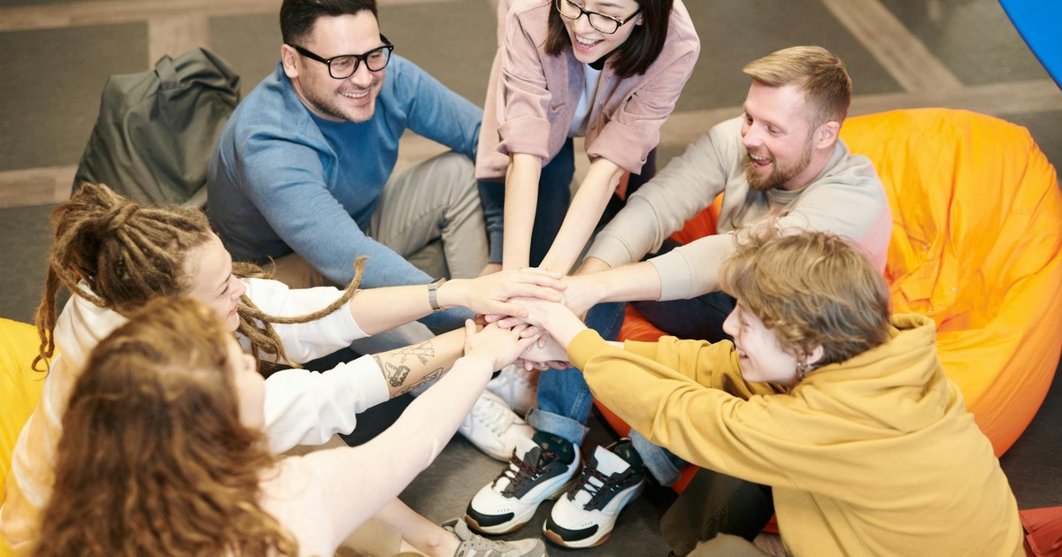 A diverse group of adults sit indoors, joyfully stacking hands together, symbolizing teamwork and camaraderie.
