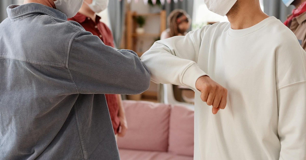 Two men bump elbows while wearing masks indoors, promoting safe greetings during COVID-19.