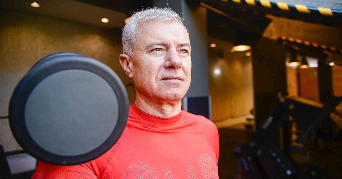 Elderly man in a gym wearing a red shirt, lifting a dumbbell, showcasing active lifestyle.