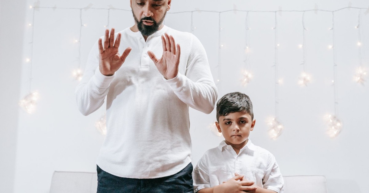 A father and son in white shirts are praying together indoors, surrounded by warm lighting.