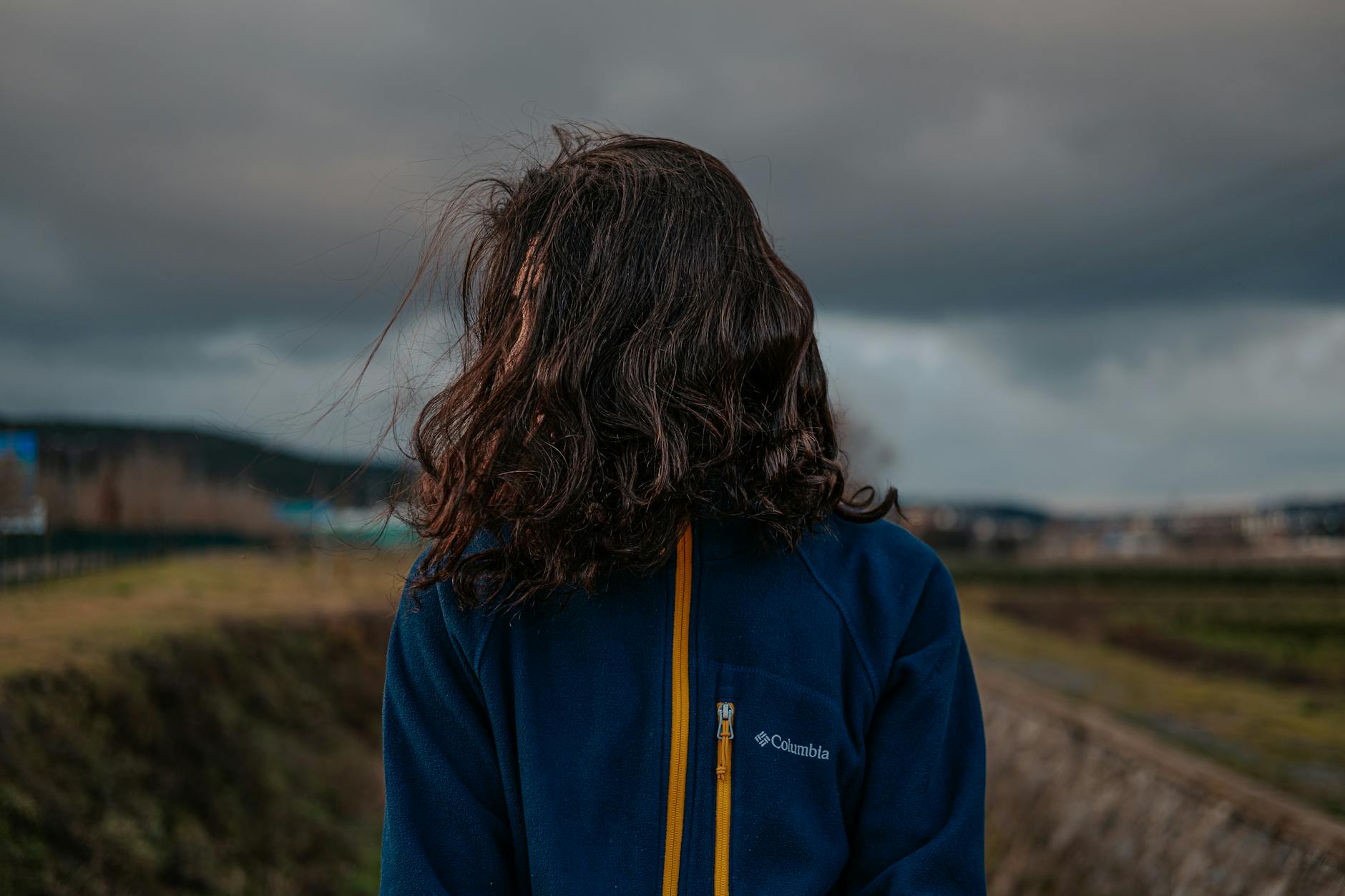 A woman stands outdoors wearing a jacket, facing away, with storm clouds overhead.